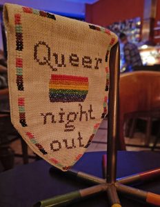 Ein weißer Fünfeckwimpel, auf dem mehrere Dinge gestickt sind: Eine Pride-Flagge, eine Bordüre mit tans- und BIPoC-Farben und die Worte Queer Night Out.