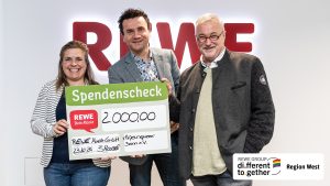 Eine Frau uns zwei Männer stahen vor einem Rewe-Logo. Sie halten vor ihnen einen großen Spendenscheck mit der Summe Zweitausend Euro.